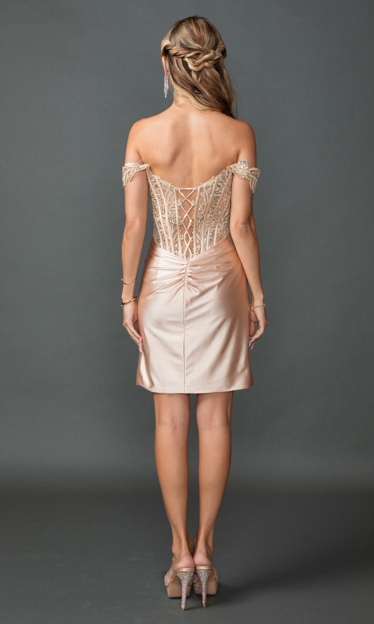 A woman stands with her back to the camera in the Short Prom Dress: Dancing Queen 3429, showcasing its off-the-shoulder lace corset bodice and fitted skirt, paired with matching heels against a plain gray background.